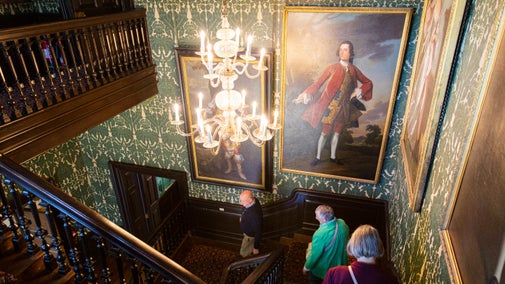 A man and two women walking down a grand staircase with art on the walls and chandelier